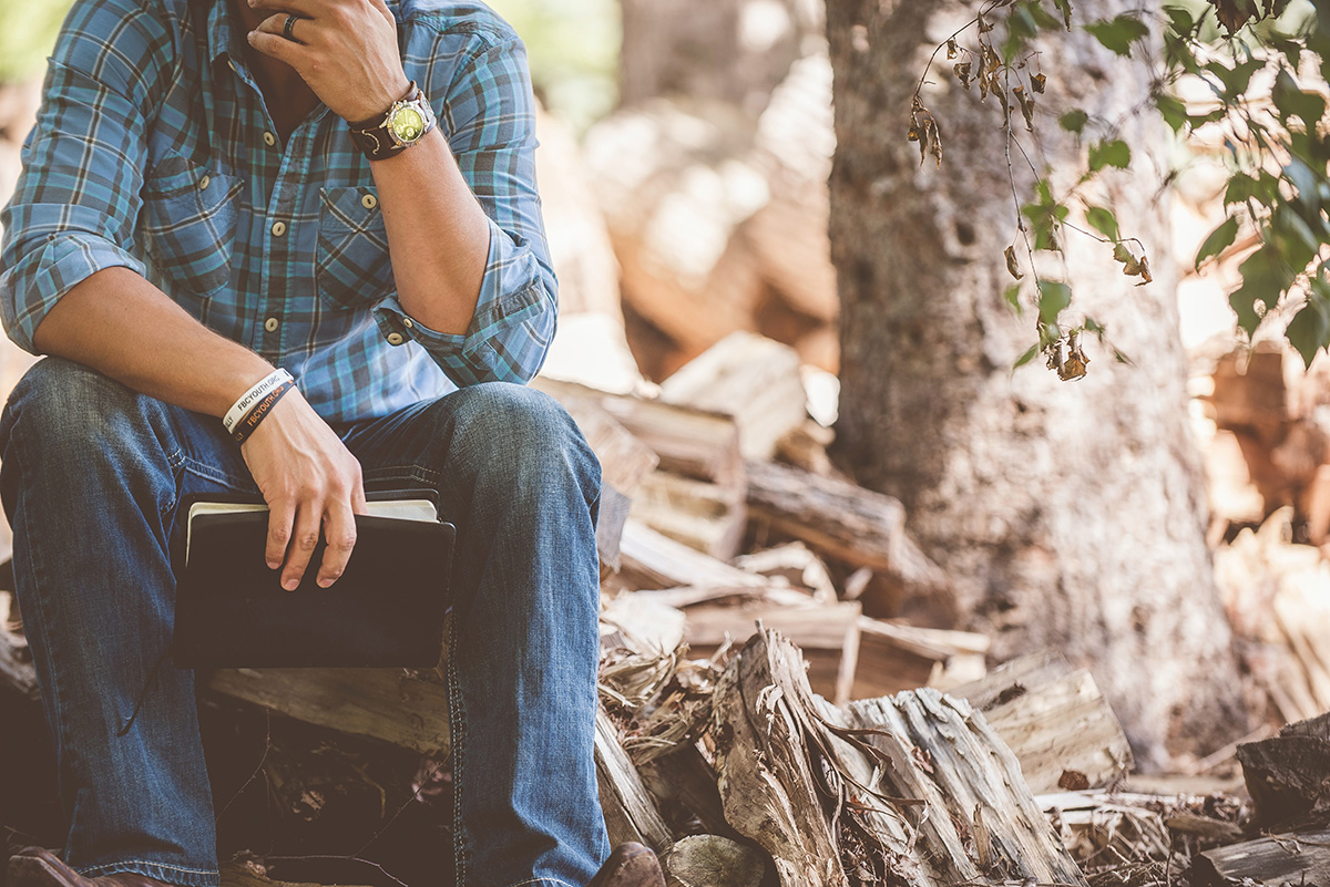 man with plaid shirt and jeans and Bible sitting in the forest thinking
